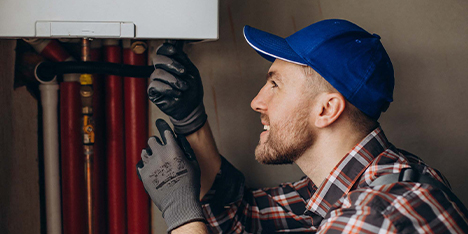 Handwerker mit grauen Handschuhen, blauer Mütze, kariertem Hemd arbeitet am Heizgerät, dunkle Wand im Hintergrund